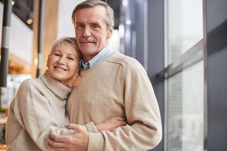 Portrait of happy beautiful senior couple in beige sweaters standing at window and cuddling each otherの写真素材