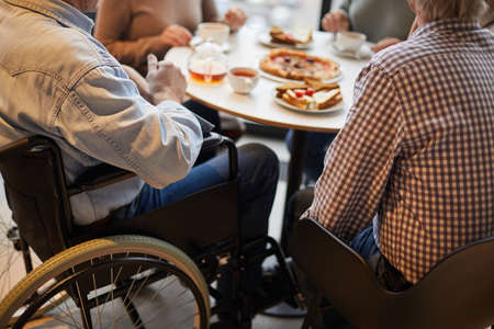 Close-up of unrecognizable handicapped man in wheelchair surrounded by friends sitting at table and chatting with friendsの写真素材