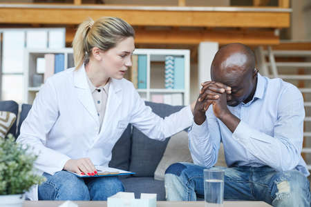 Young female psychologist in white coat sitting on sofa and touching shoulder of Black male patient while consoling him at therapyの写真素材