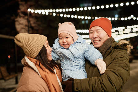 Horizontal medium shot of joyful Asian couple and their lovely toddler daughter spending time outdoors on winter eveningの写真素材