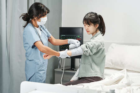 Horizontal shot of medical worker wearing mask on face measuring blood pressure of woman in emergency roomの写真素材