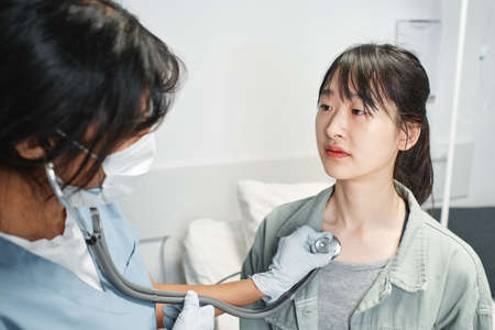 Doctor wearing protective mask on face using stethoscope listening to inner sounds of young Asian patientの写真素材