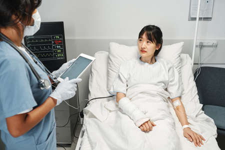 Unrecognizable doctor wearing mask holding digital tablet working with young Asian woman in emergency roomの写真素材