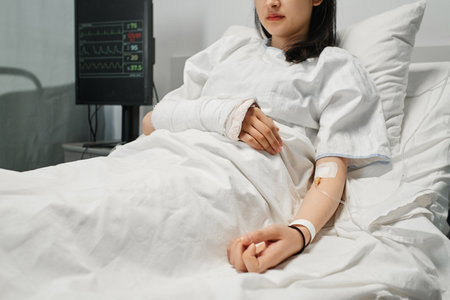 Horizontal shot of young Asian woman with arm in cast lying on hospital bed in emergency room getting intravenous therapyの写真素材