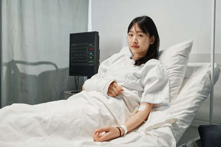 Horizontal medium shot of young Asian woman with arm broken after accident lying on hospital bed in emergency room getting IV therapyの写真素材