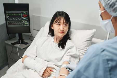 Horizontal over-the-shoulder shot of young Asian woman lying on hospital bed getting IV therapy in emergency roomの写真素材