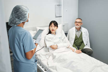 Unrecognizable doctor holding digital tablet talking to young Asian patient and her relative in emergency roomの写真素材