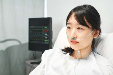 Medium close-up portrait of young Asian woman wearing nasal cannula sitting on hospital bed looking awayの写真素材