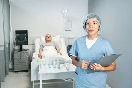 Horizontal medium portrait of woman working in hospital holding clipboard looking at camera, her patient lying in bed on backgroundの写真素材