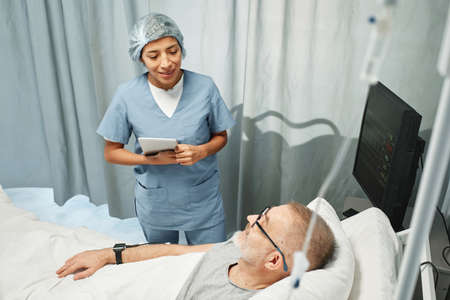 Horizontal high angle shot of young adult woman working in hospital holding digital tablet telling something to mature man in bedの写真素材