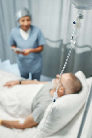 Selective focus on foreground shot of mature man getting IV therapy in modern hospital talking to his doctorの写真素材