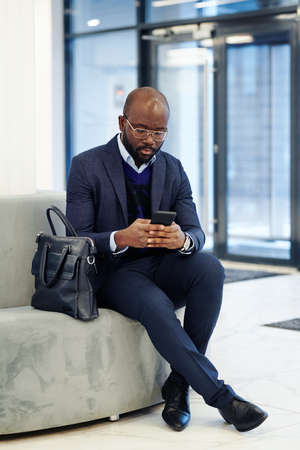African businessman typing a message on his mobile phone while sitting on the couch in the lobby and waiting for the meetingの写真素材