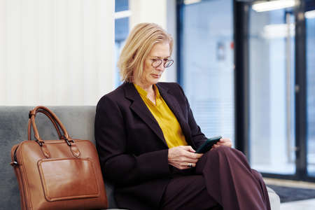 Mature businesswoman sitting on the couch and working online on mobile phone while waiting for the meeting in the lobbyの写真素材