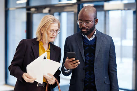 Mature female lawyer with documents discussing the deal with her colleague while he using his mobile phone before the meetingの写真素材