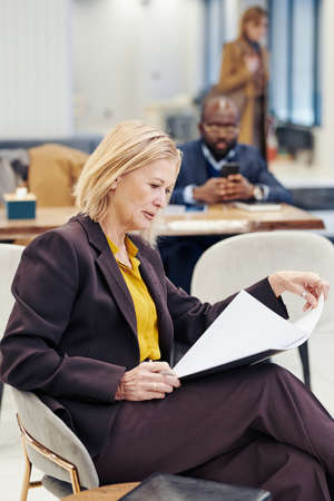 Mature female lawyer in suit sitting on chair and examining the document before meeting with her colleagues in the backgroundの写真素材
