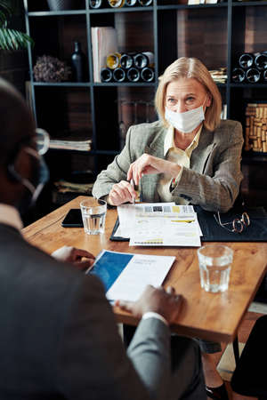 Mature lawyer in mask sitting at the table with documents and discussing law deal with her colleague during their meeting in the restaurantの写真素材