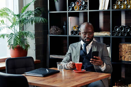 African businessman in suit sitting at the table with cup of coffee at the restaurant and typing a message on mobile phoneの写真素材