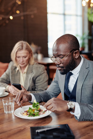 African businessman enjoying his salad sitting at the table at restaurant together with his colleague in the backgroundの写真素材
