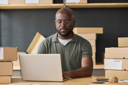 African warehouse worker concentrating on his online work on laptop while sitting at tableの写真素材