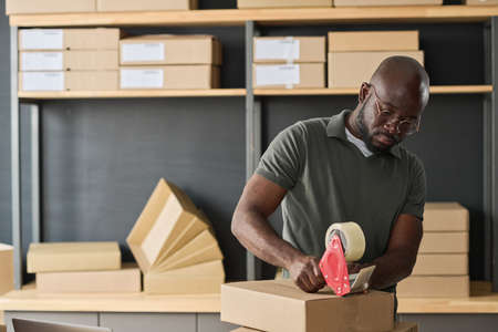 African warehouse worker in eyeglasses packing cardboard boxes with adhesive tape before shippingの写真素材