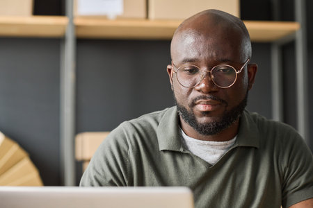 African serious man in eyeglasses working online using laptop in warehouse