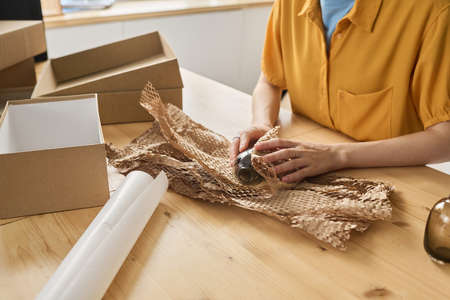 Close-up of woman wrapping bottle in paper and packing it in cardboard box at table for deliveryの写真素材