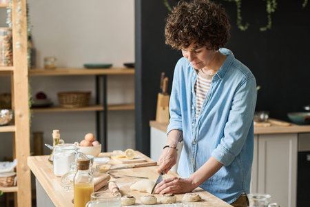 Young woman cutting dough for little pieces to bake homemade buns at table in kitchen