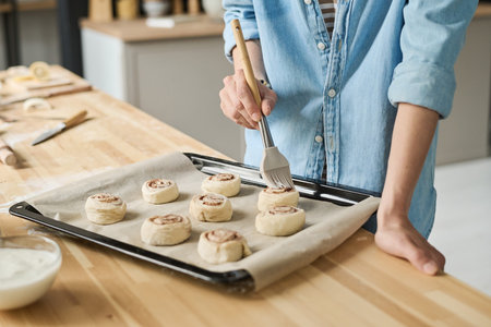 Close-up of young woman using brush to oil buns on tray before baking