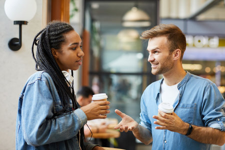 Content handsome young man in denim shirt holding coffee and talking to Afro-American girl in coffee shopの写真素材