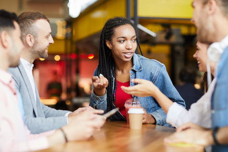 Group of content young multi-ethnic students in casual clothing sitting at table and drinking beverages while chatting in cafeの写真素材