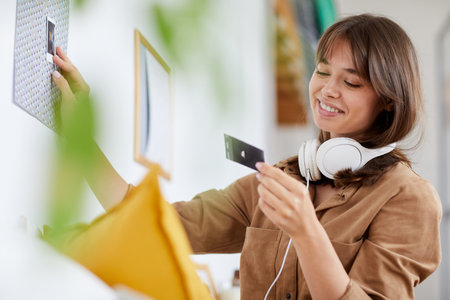 Smiling attractive young woman with brown hair looking at instant photo while decorating room with photographsの写真素材
