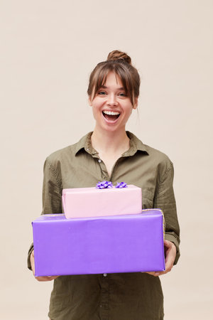 Thrilled beautiful young lady with bangs and hair bun standing against beige wall and holding stack of gift boxesの写真素材