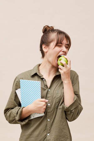 Young female student with hair bun wearing khaki shirt holding workbooks and eating green apple