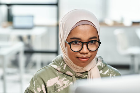 Young confident female IT support manager in eyeglasses and hijab sitting in front of computer monitor and networking in officeの写真素材