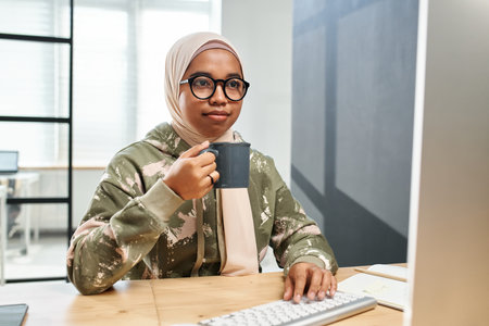 Young serious IT engineer in hijab, casualwear and eyeglasses having coffee in front of computer monitor by her workplace in officeの写真素材
