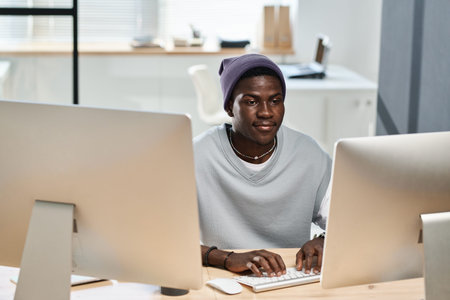 Young black man in casualwear working over new software while sitting in front of computer monitors...