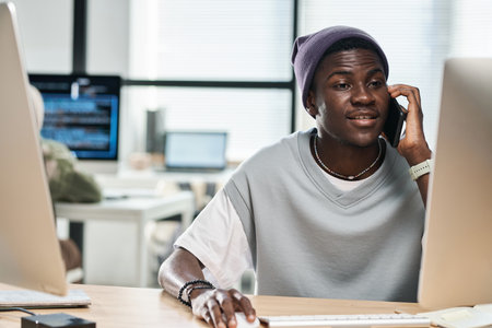 Young confident African American software developer consulting client on mobile phone while sitting in front of computer monitor in officeの写真素材