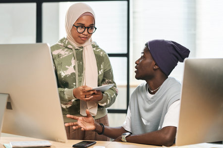 Two young multicultural programmers in casualwear having discussion of new project points while Muslim woman listening to male colleagueの写真素材