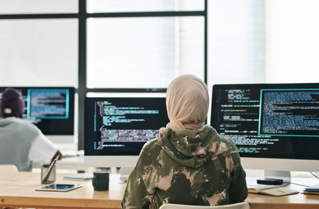 Rear view of young Muslim woman in hijab and casualwear sitting by workplace in front of computer screens with coded dataの写真素材