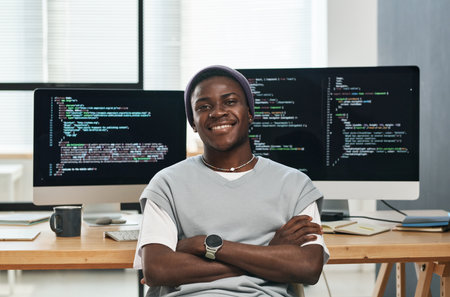 Happy young successful IT engineer in casualwear looking at you while sitting by workplace against computer screens with coded dataの写真素材