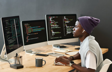 Young serious IT engineer in casualwear looking at coded data on computer screens while typing on keyboard by workplaceの写真素材