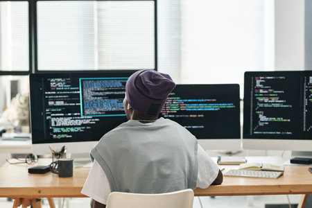 Back view of young software developer sitting by workplace in front of three computer monitors with...
