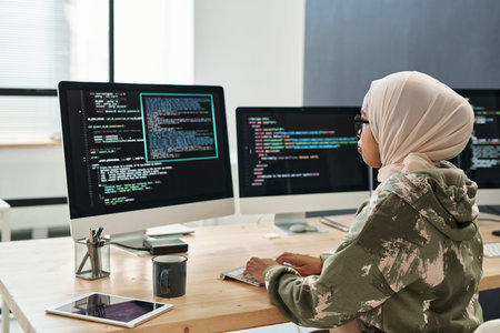 Side view of young Muslim businesswoman networking in front of computer screen with coded...