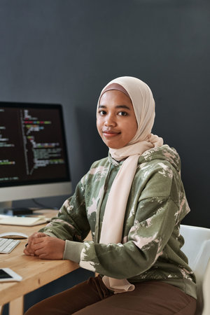 Young successful Muslim businesswoman in casualwear sitting by workplace against computer screen...