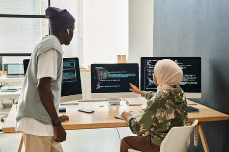 Young confident Muslim woman in hijab making presentation of decoded data on computer screen to African American male colleagueの写真素材