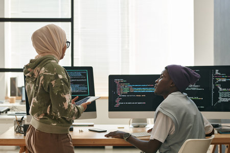 Two young intercultural IT engineers discussing ways of decoding data while Muslim woman in hijab standing in front of computer screensの写真素材