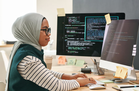 Young Muslim businesswoman in hijab looking at computer screen while typing and decoding data in front of monitor in officeの写真素材