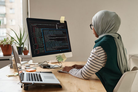 Young Muslim businesswoman or intern looking at computer screen while typing and decoding data or developing software by workplaceの写真素材