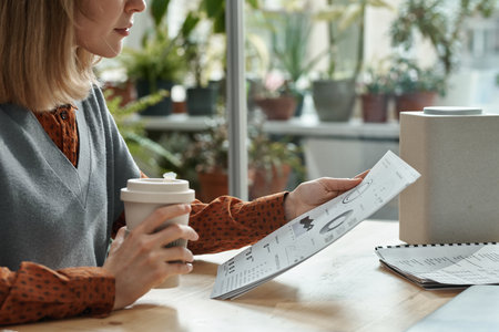 Close-up of young woman drinking coffee and reading document with graphs while sitting at table at officeの写真素材