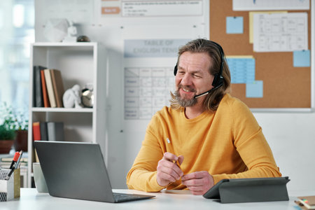 Mature teacher in headphones sitting at his workplace with laptop and talking to his students onlineの写真素材